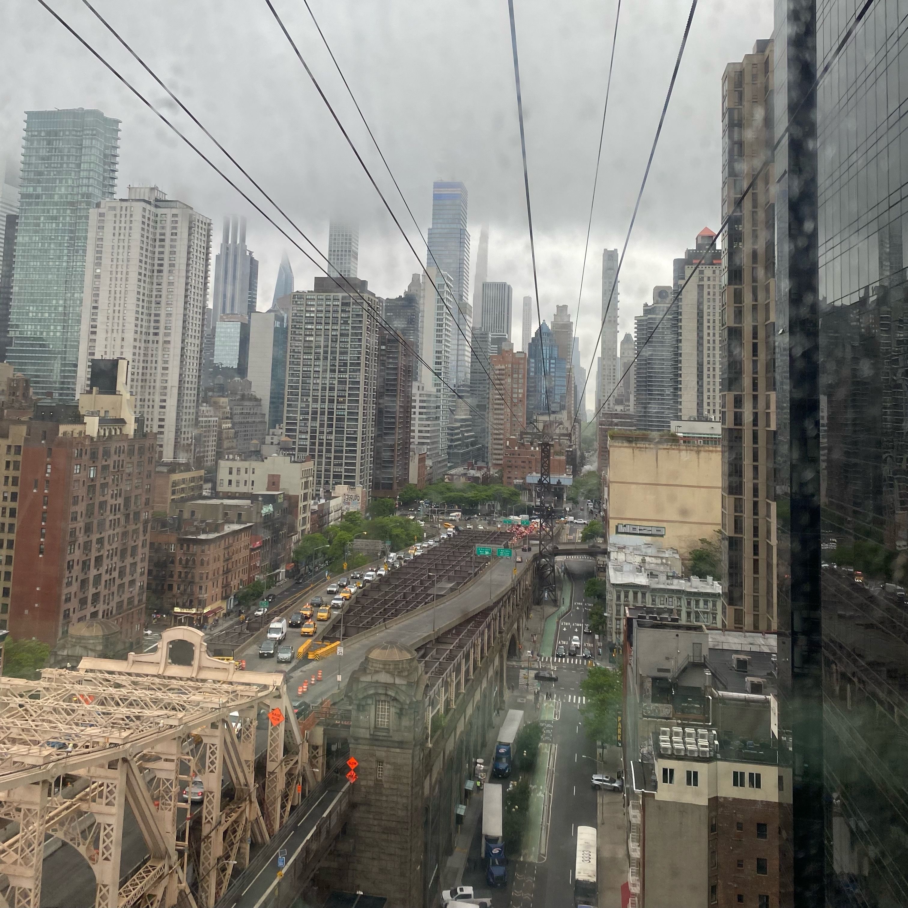 An aerial view of a bridge with several cars streaming from streets filled with skyscrapers. The sky is overcast and streaked with lines leading towards a tram tower.