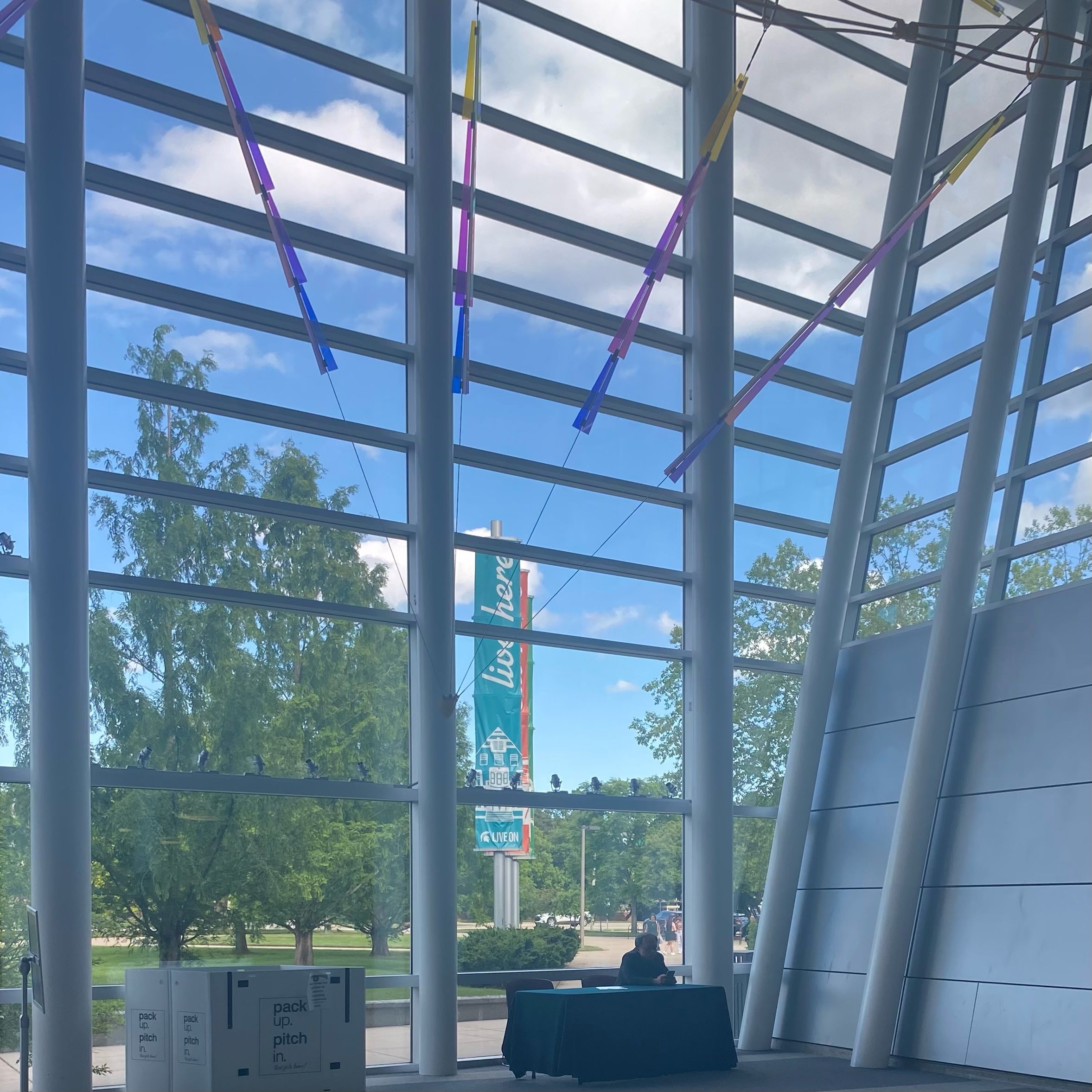 A glass building wall with a grid structure. There is a view of several trees outside. A man is sitting at a table at the bottom of the wall.