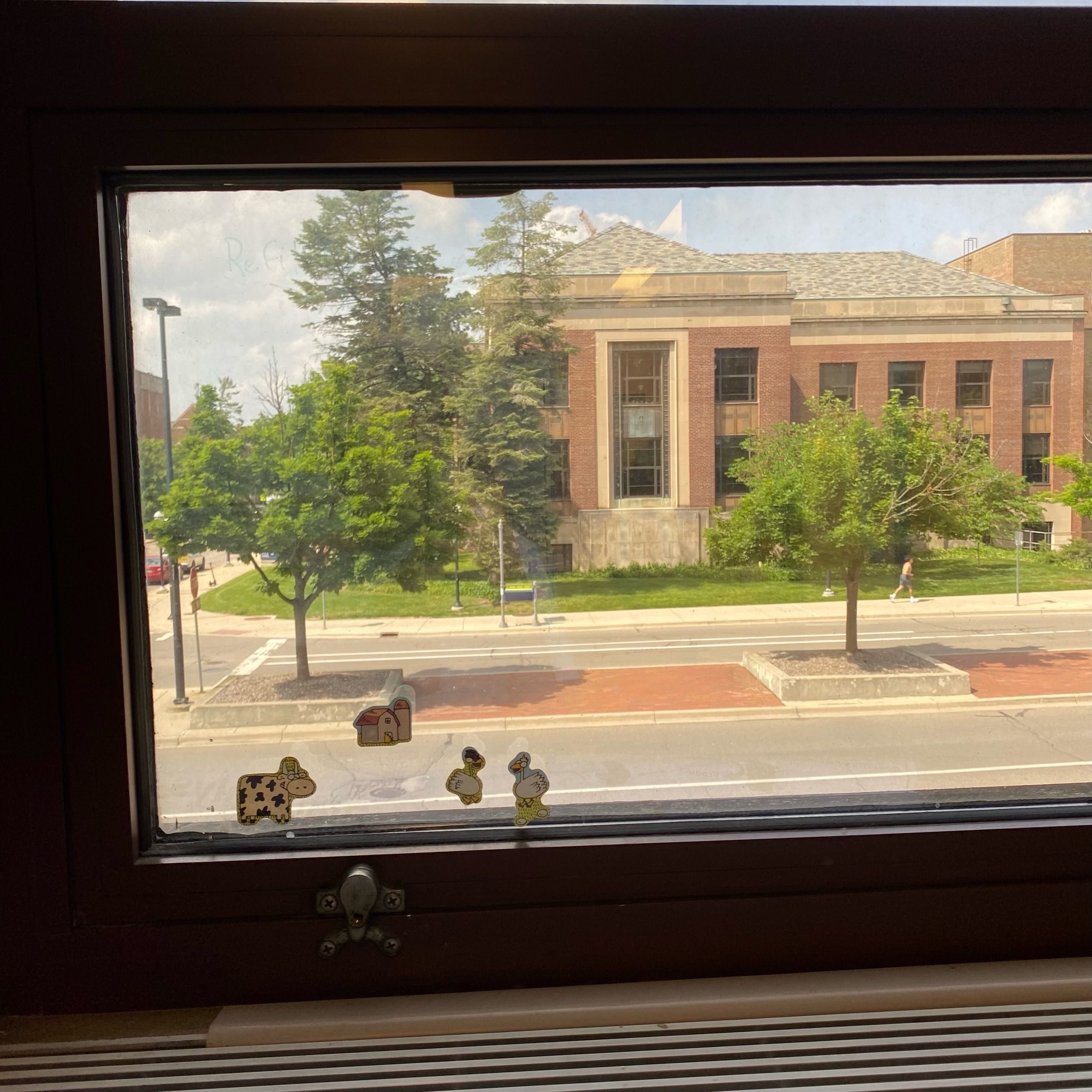 A window with a view of a brick building and several trees. The window has farm stickers on it.