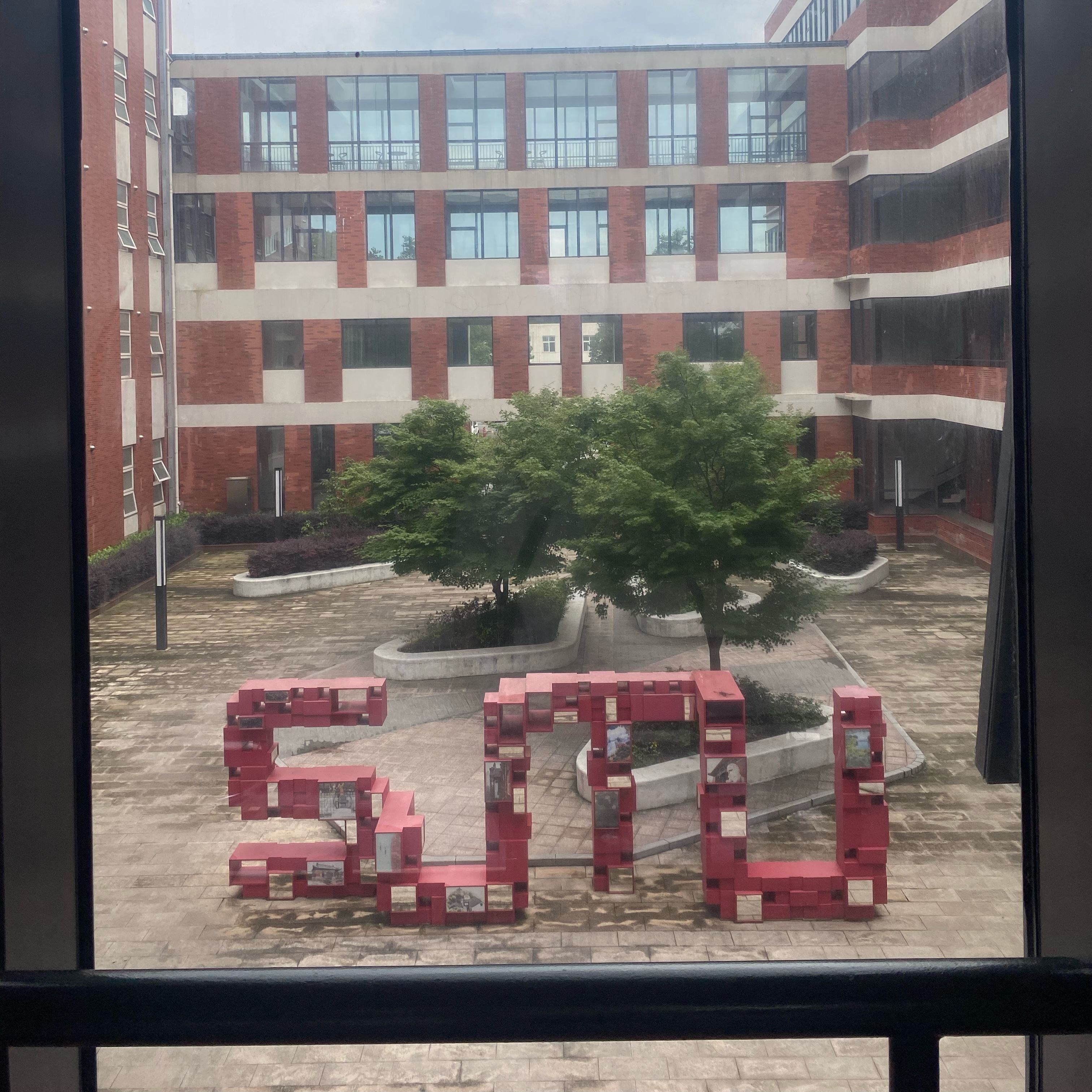 A window with a view of red 'SJTU' block letters in a courtyard.