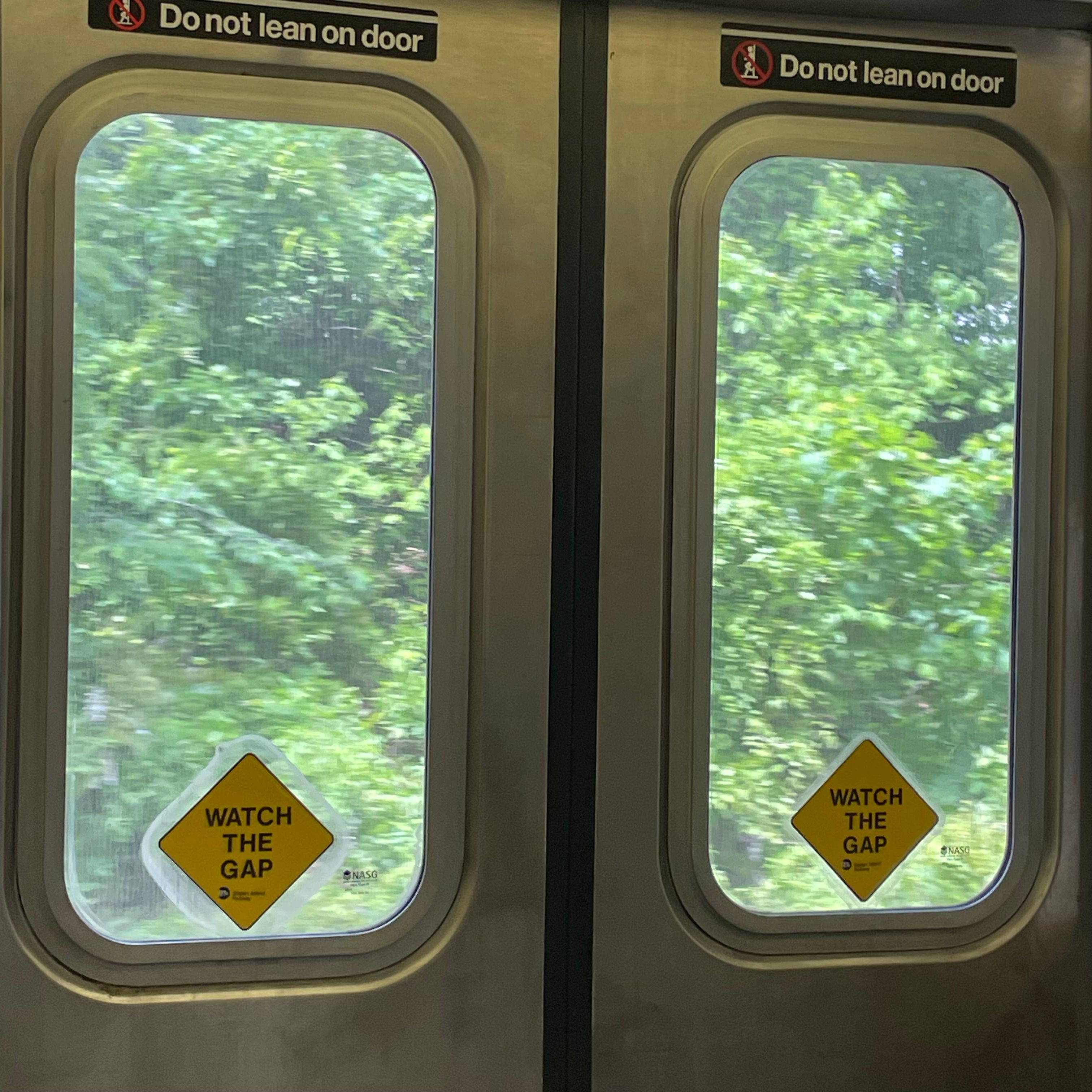 A New York City subway door. The doors are labeled with 'Do not lean on door' and 'Watch the gap' stickers. The doors windows have a view of green trees.