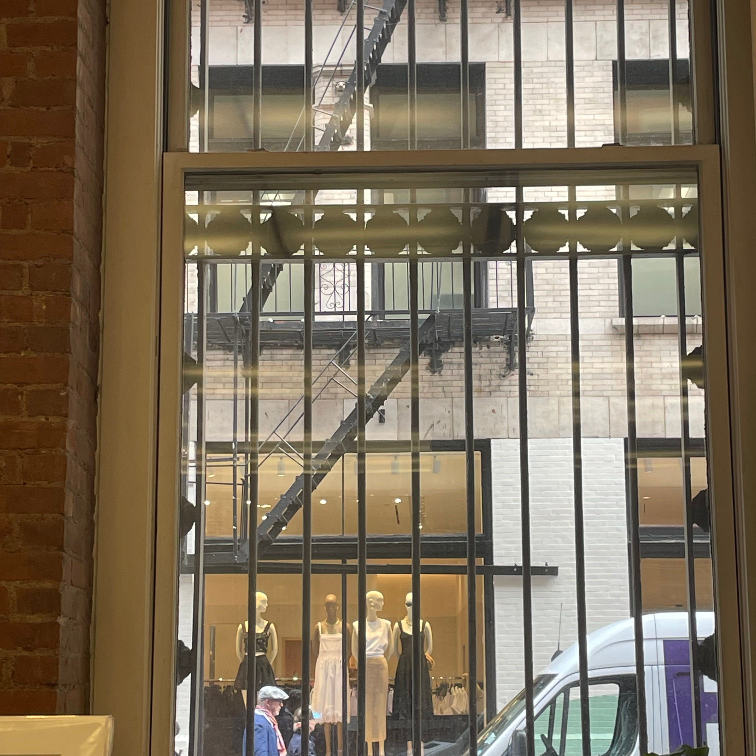 A window with a view of four mannequins behind another building's window. There is a fire escape above the window. A man walks past the window towards a FedEx truck.