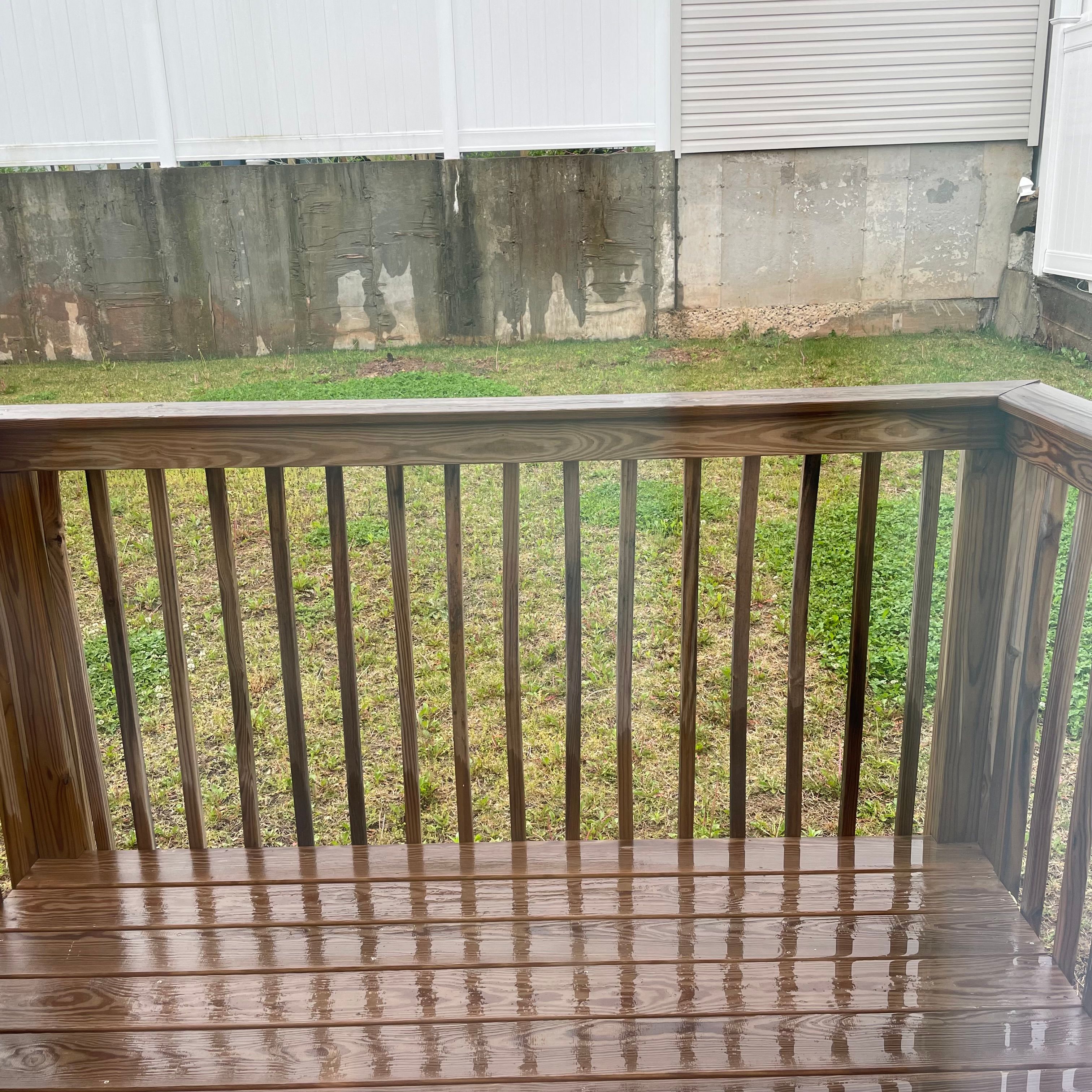 A wooden porch in front of a patch of grass and a concrete wall and white fencing. The porch is wet with a reflective quality.