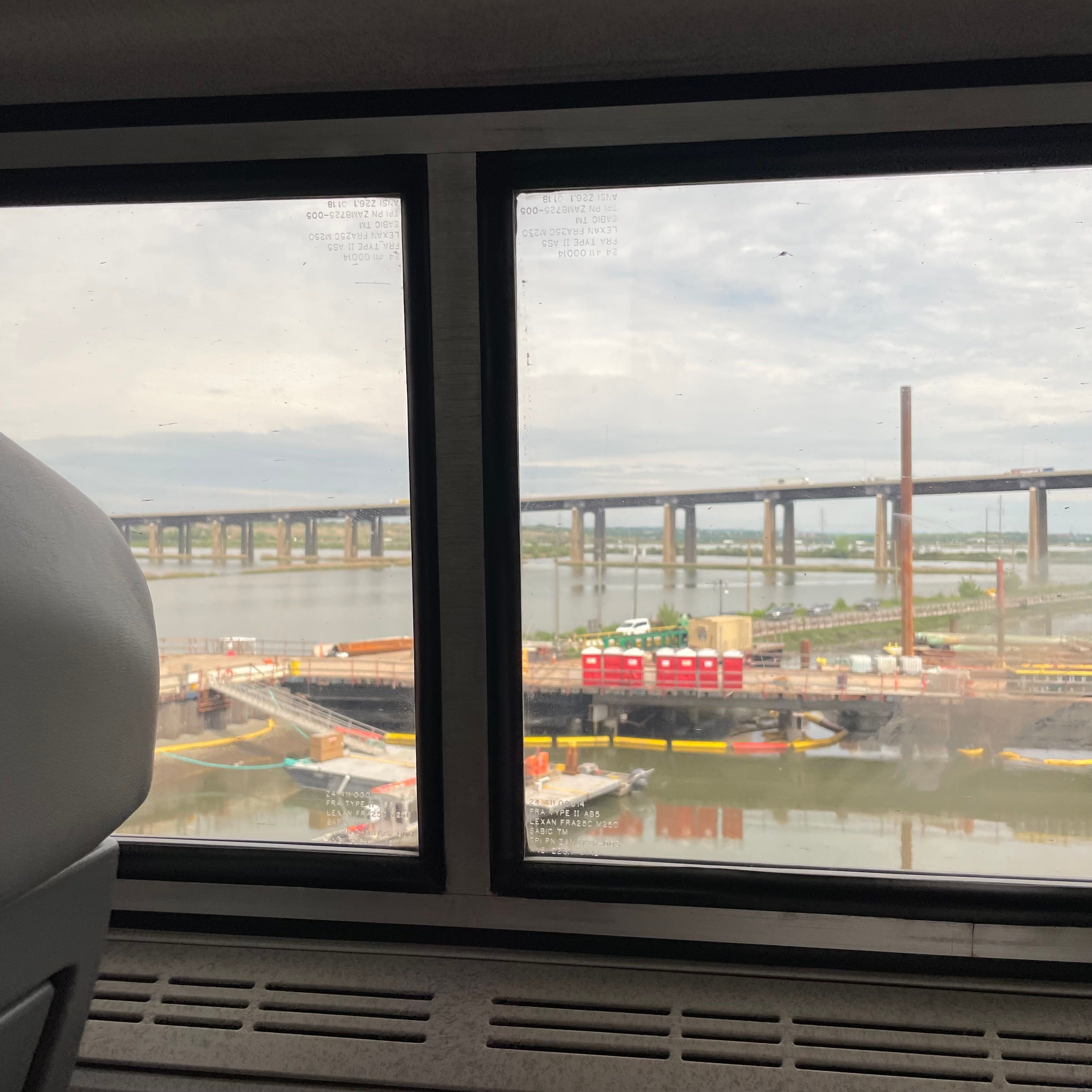 A train window with a view of a bridge going over a river. There is a pier in the foreground with various containers and a row of red portable toilets.