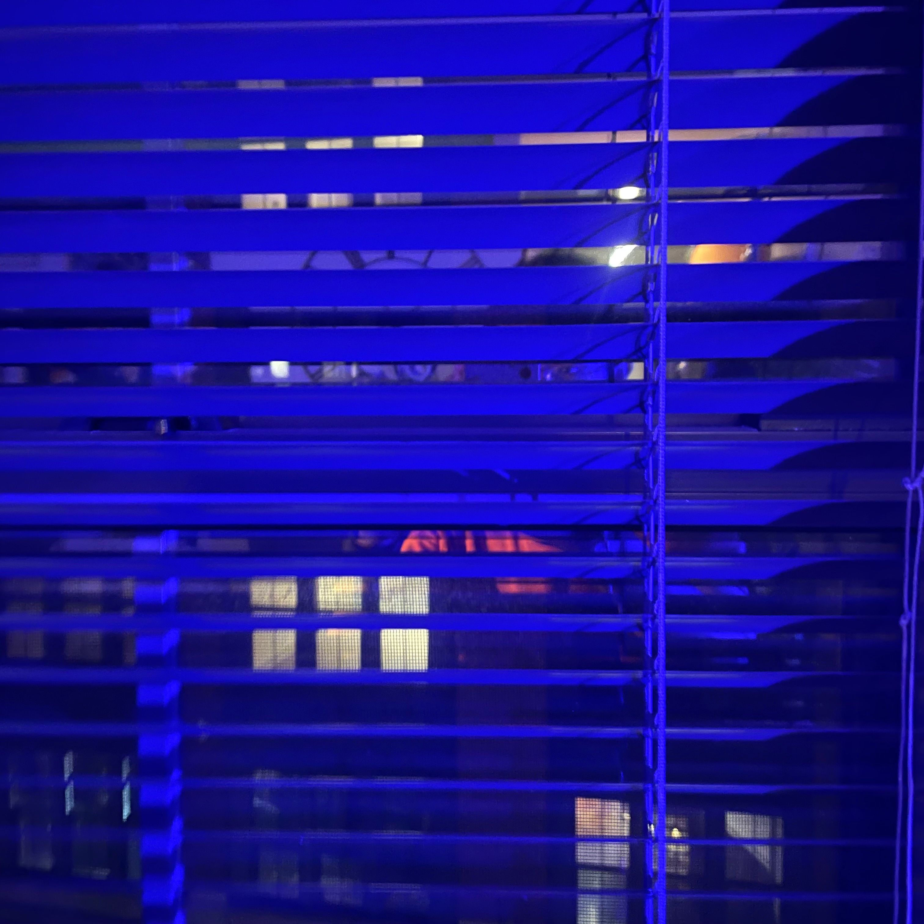 A window with blinds tinted dark blue from an ambient light. The window has a view of several other windows with yellow light streaming out of them.