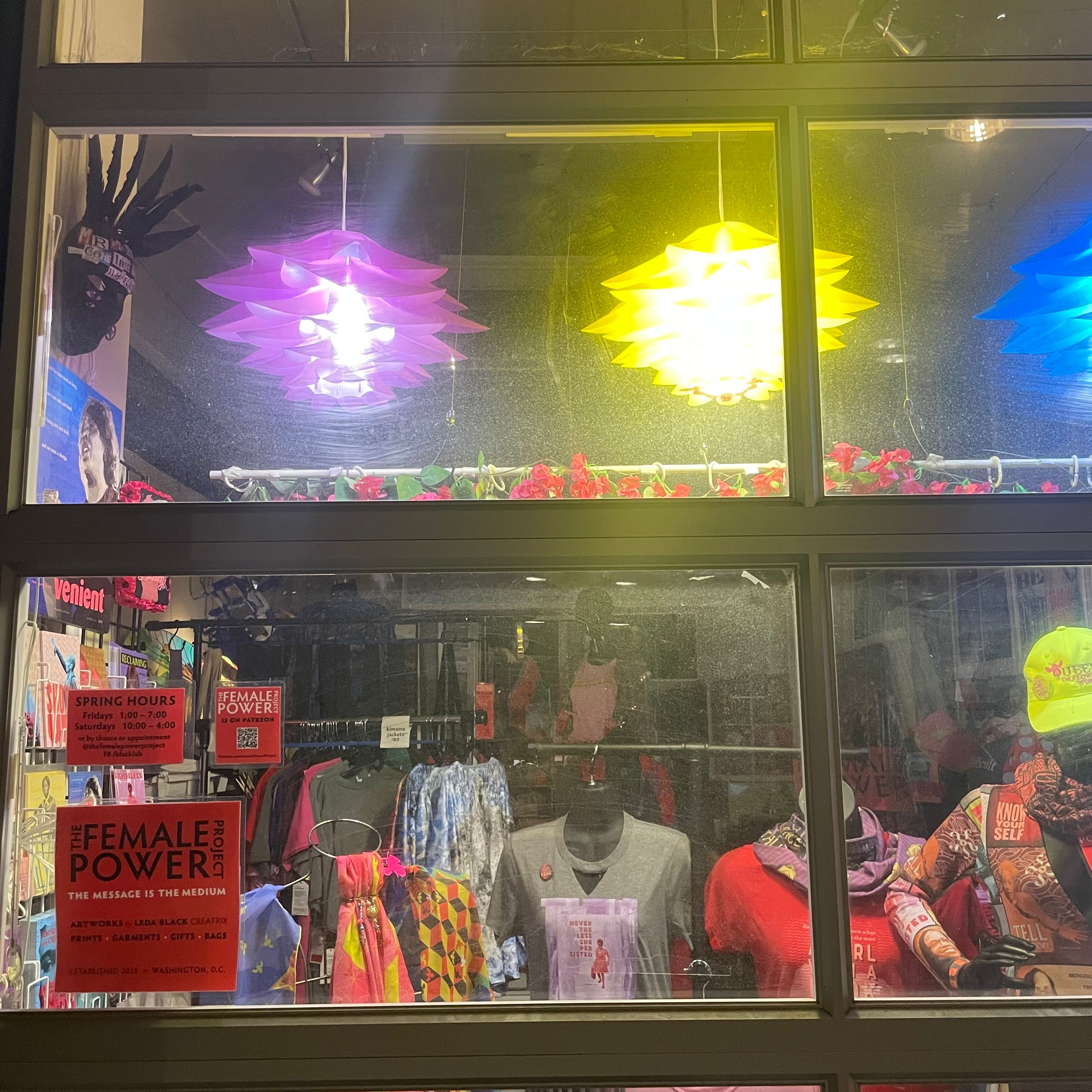 A window with a view of several mannequins wearing shirts, rows of clothing, and colorful lighting. There is a red sticker on the window surface that says 'The Female Power Project.'