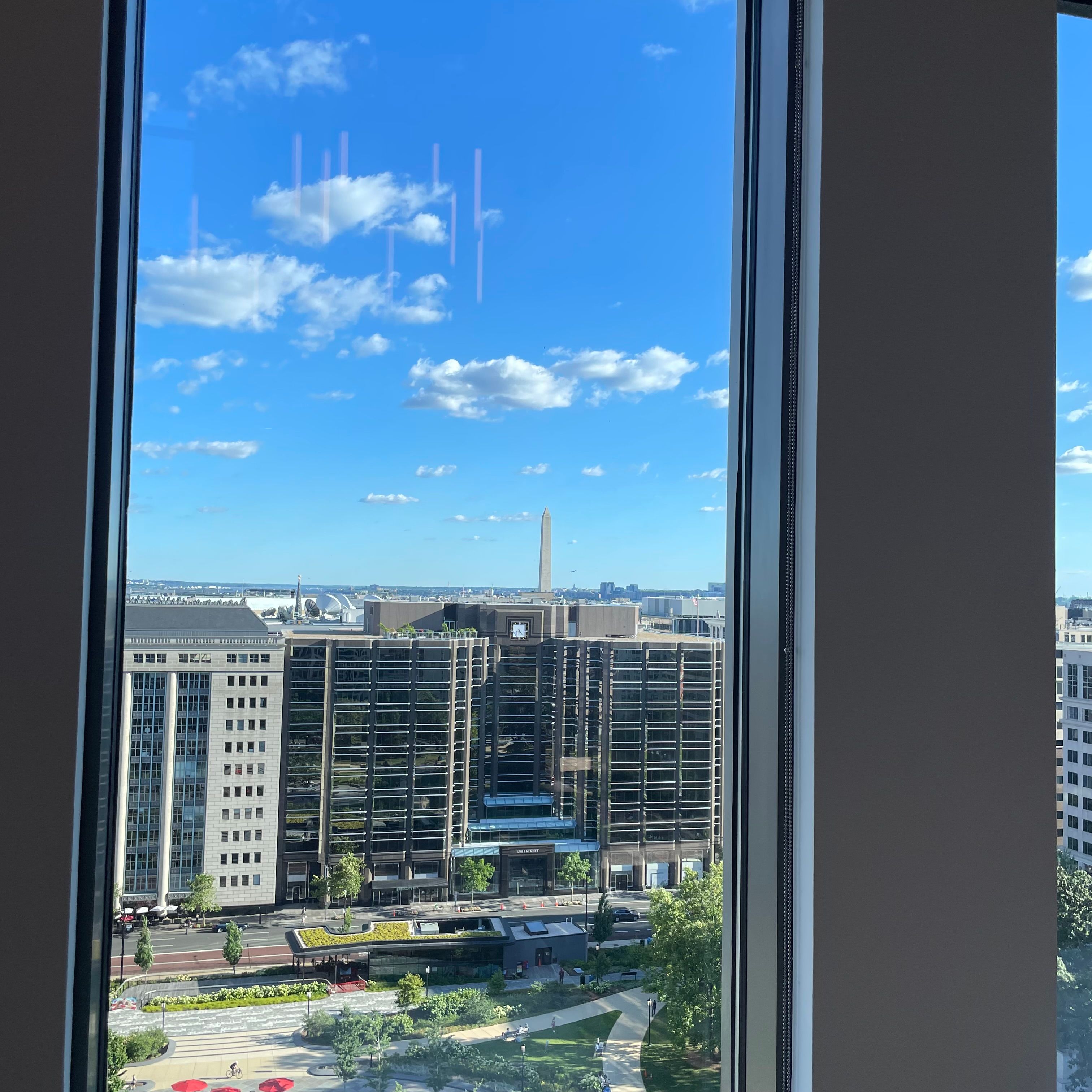 A window with a view of the Washington Monument in the background. It is surrounded by other buildings in the foreground. The sky is largely clear with a few clouds and reflective streaks from the window.