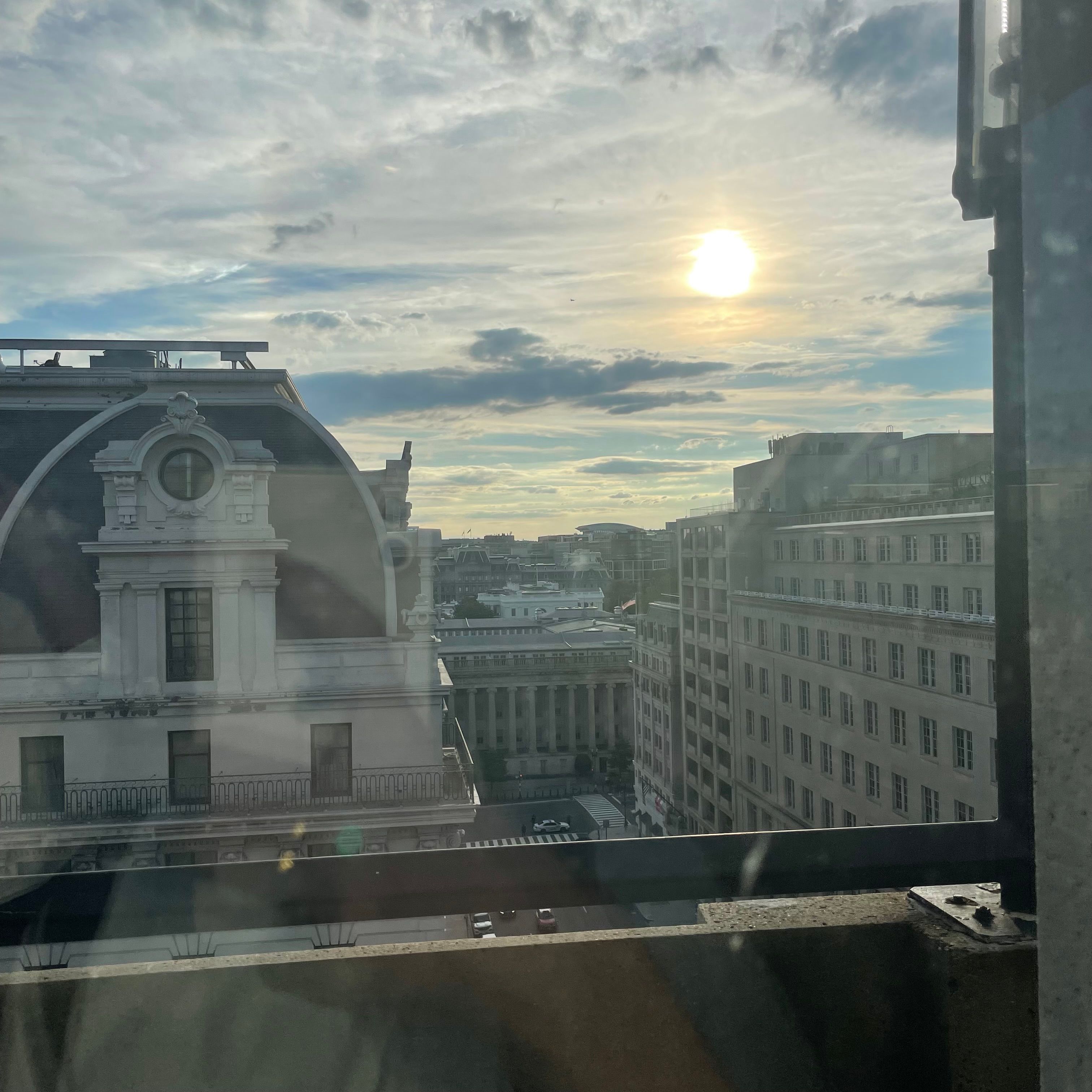 A window with a slight reflection of me. There is a view of a partly cloudly sky with a yellow sun peaking through. The White Hosue is visible in the background, surrounded by other taller buildings.