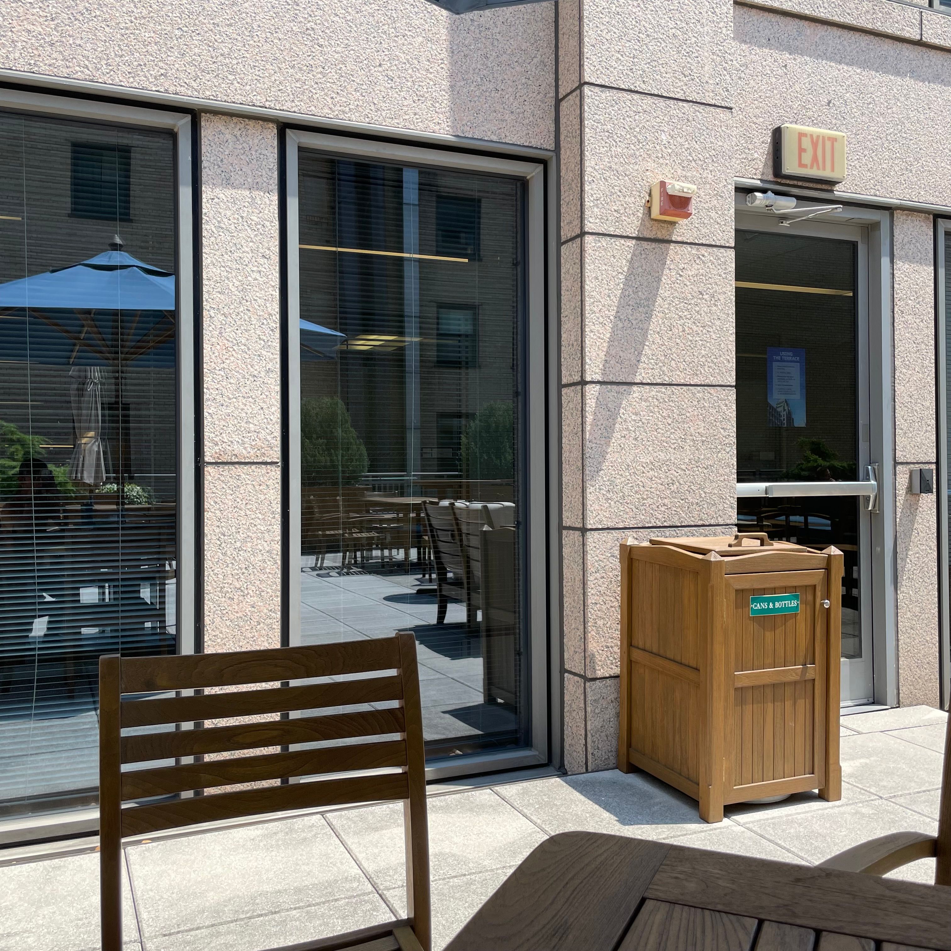 Two tall windows with a reflection of a blue table umbrella. The windows are next to a glass door with an exit sign on top of it. There is a table and chair in the foreground.